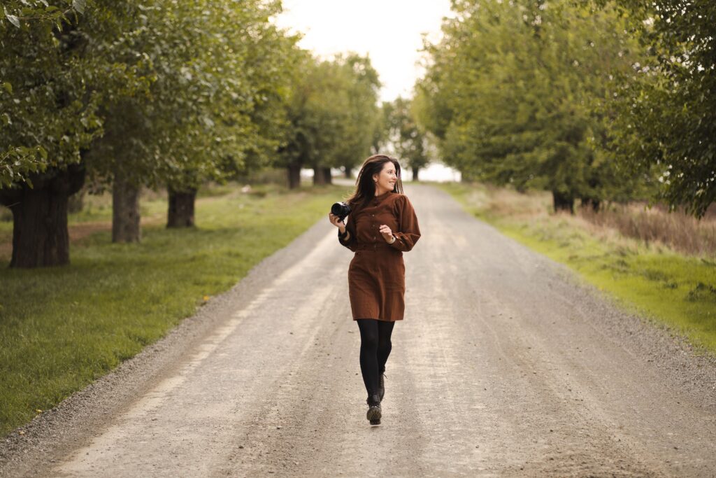 A woman photographer joyfully walks down a scenic tree-lined path in autumn.